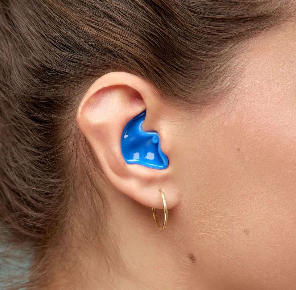 Detajlni pogled na žensko uho v katerem je ušesni čepek v modri barvi. Na ušesu se sveti zlat okrogli uhan.