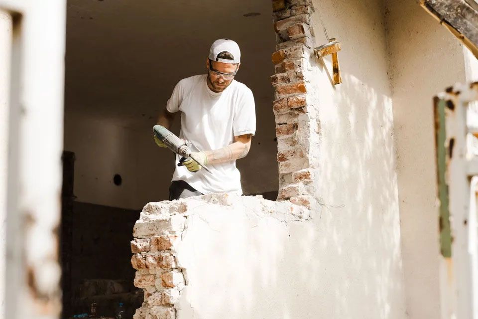 Moški v beli majici s kratkimi rokavi, zaščitnimi očali in kapo s šiltom obrnjeno nazaj, v rokah drži sveder in stoji nad na pol podrto steno. Zraven je stena v svetli barvi.