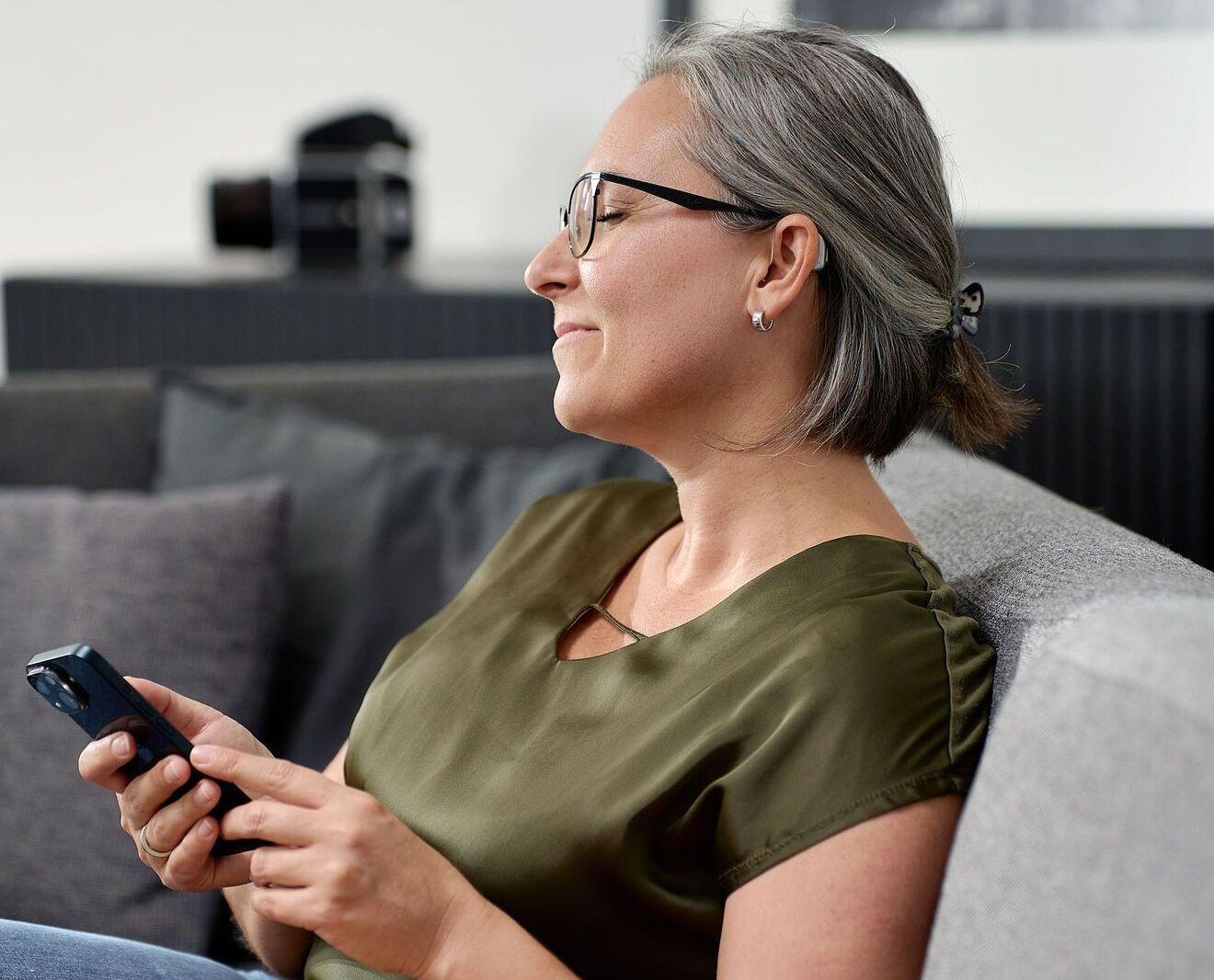 Femme assise sur un canapé, tenant un téléphone à la main, portant un appareil auditif dans l’oreille