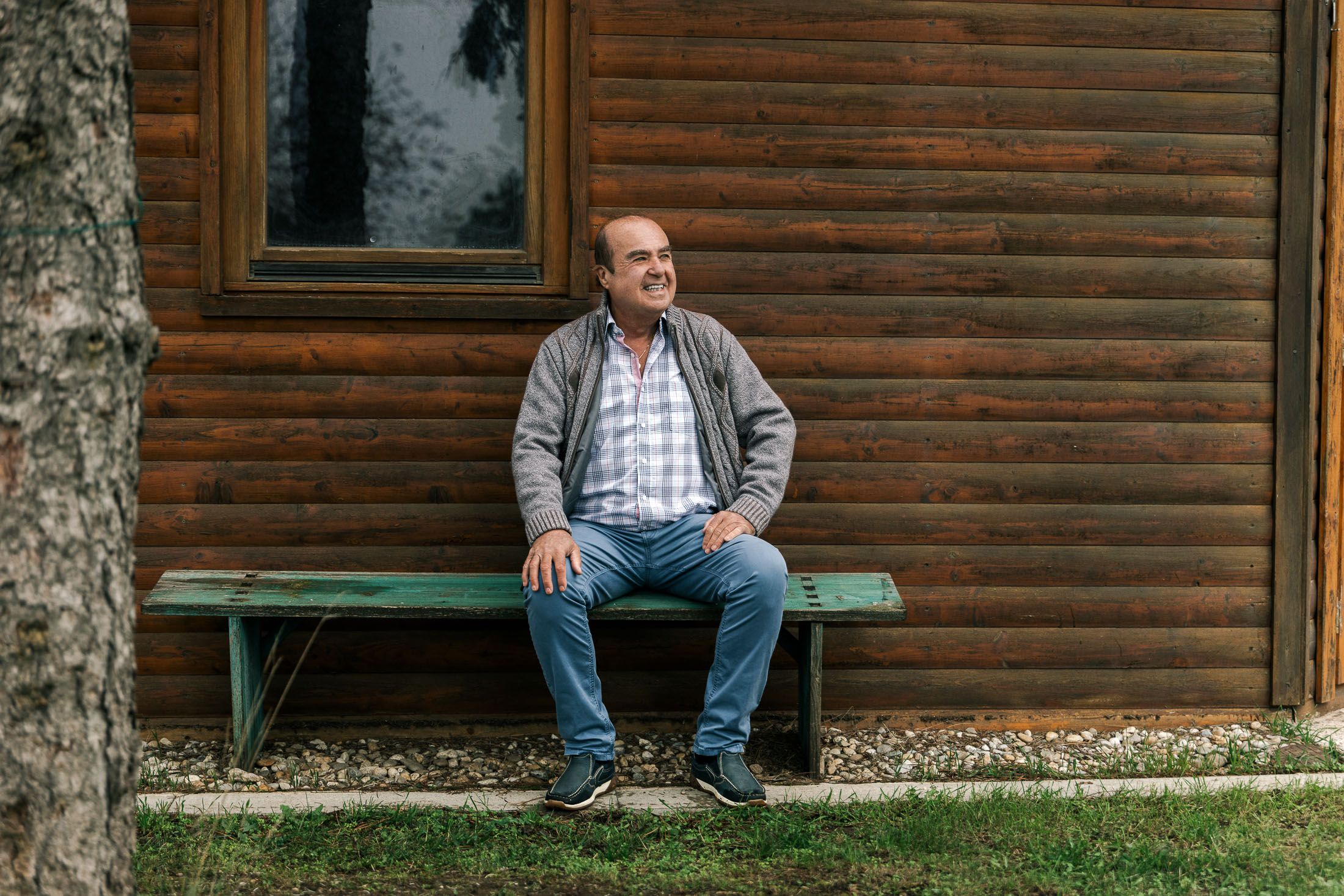 Älterer Herr sitzt auf grüner Bank vor Holzhaus. Lächelt und sieht in die Ferne.