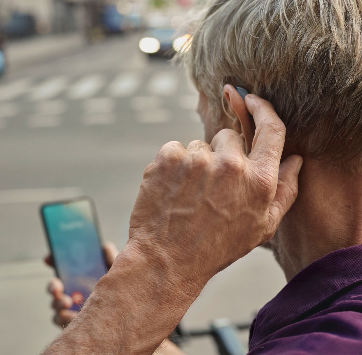 Ein Mann der Im Straßenverkehr auf sein Handy schaut und sich dabei das Hörgerät hinter dem Ohr richtet. 