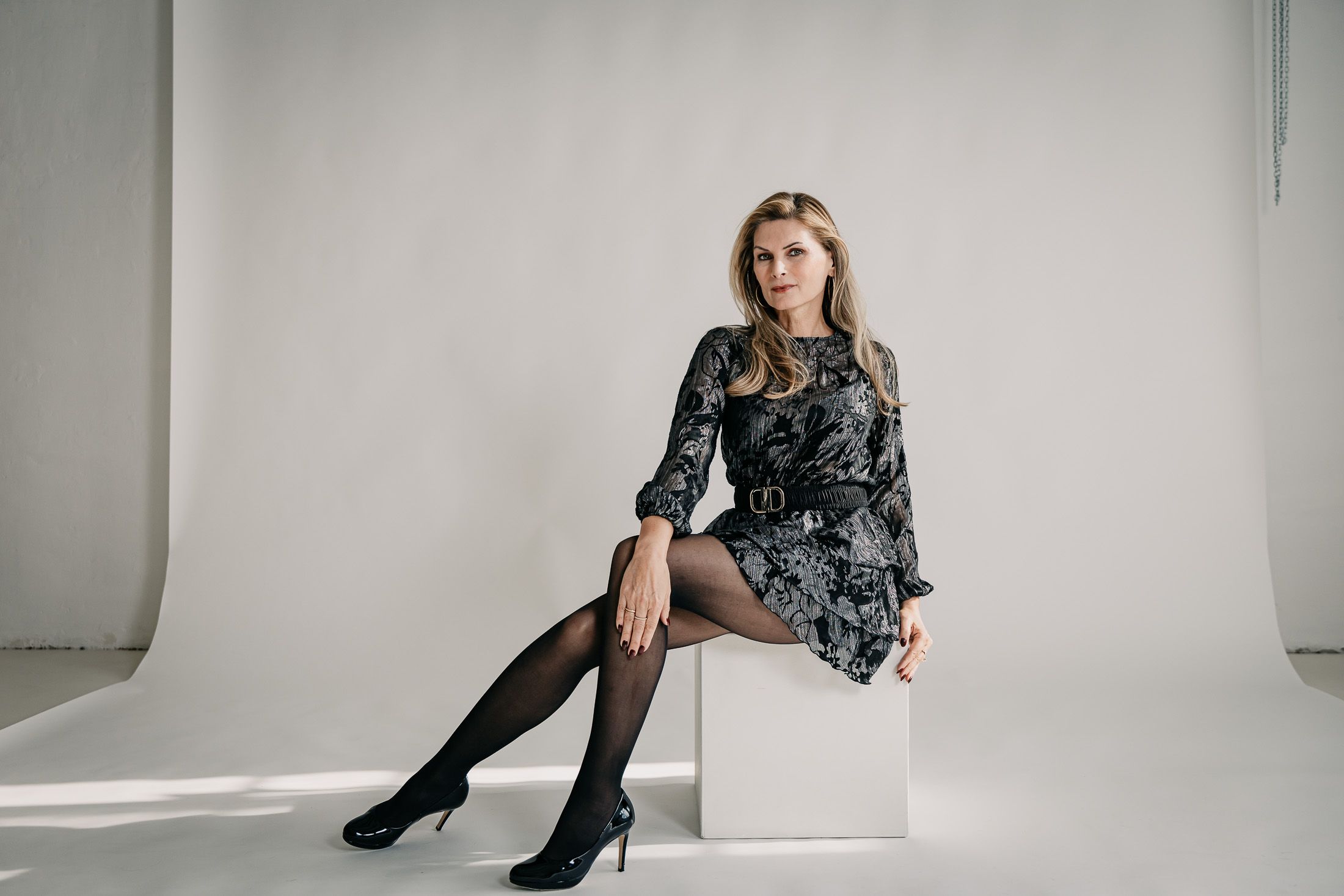 Martina Kaiser posiert auf einem weißen Sitzwürfel. Trägt weiß-silbernes Kleid, schwarze Strumpfhose und schwarze High-Heels.
