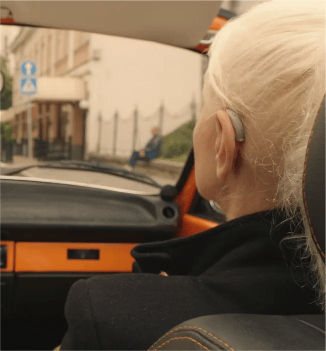 Starija žena sede kose snimljena otpozadi sedi u automobilu na mestu suvozača i nosi slušni aparat