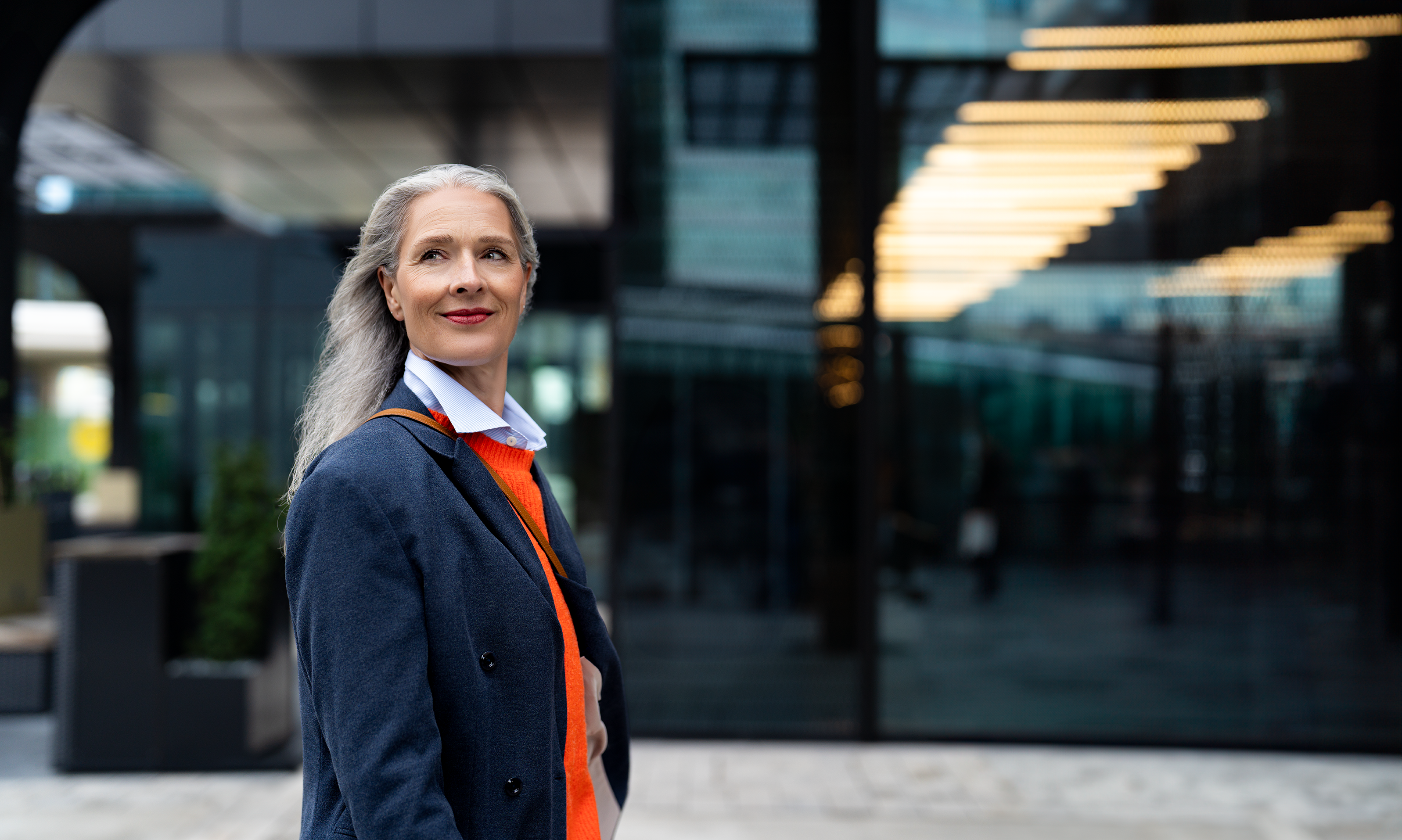 Ältere, modische Frau mit grauen, langen Haaren blickt stolz in die Ferne und lächelt.