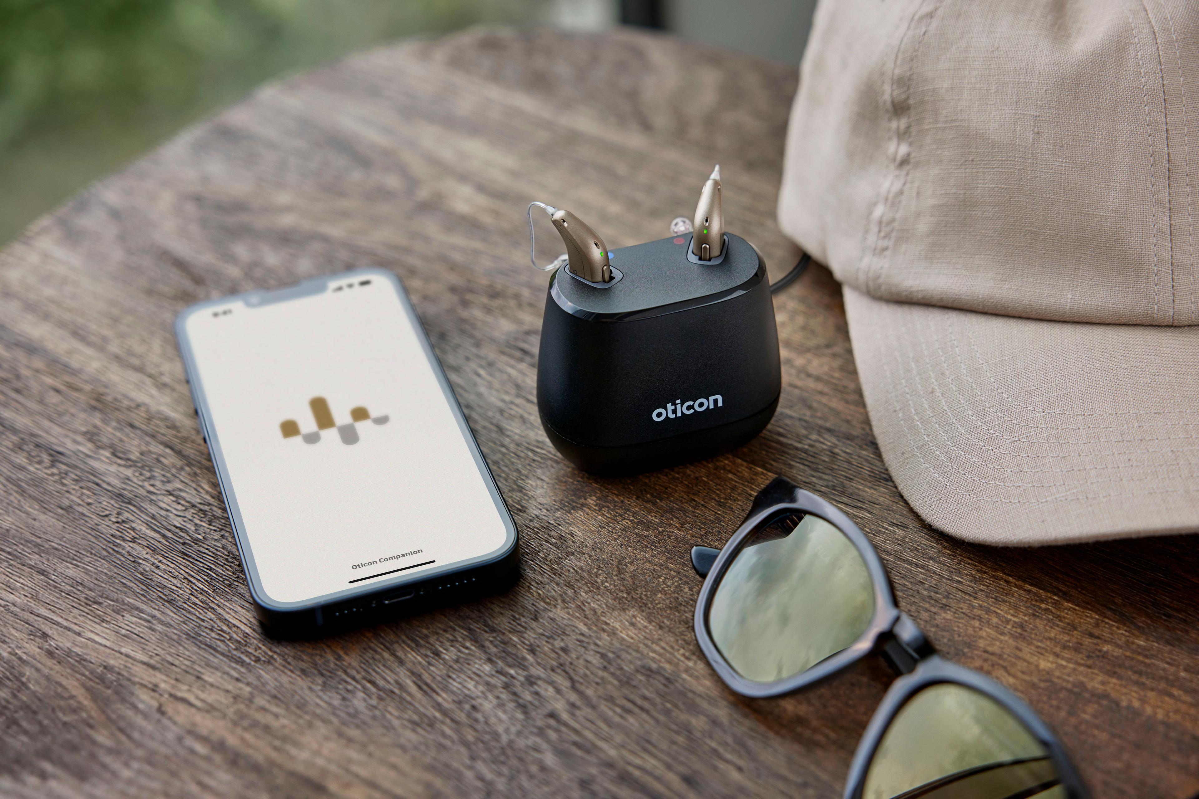 Oticon Intent im Charger, daneben Handy und Sonnenbrille