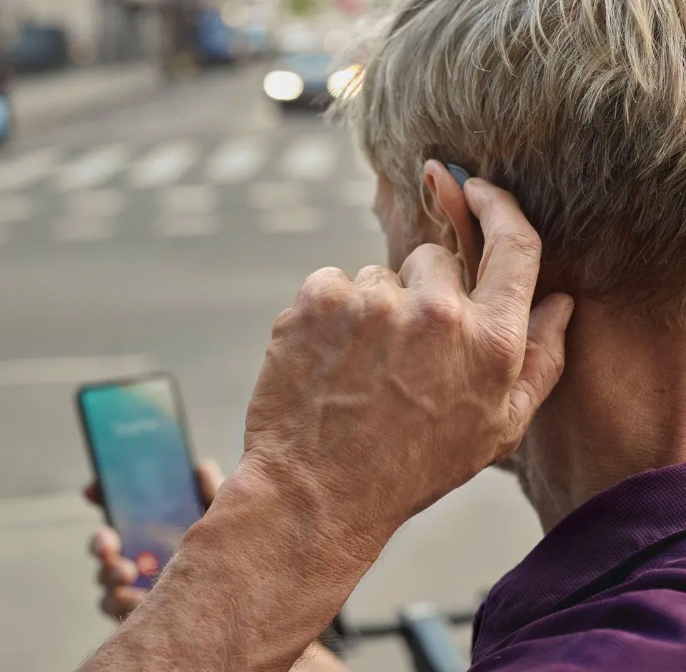 Starejši moški si z levo roko uravnava zunanji slušni aparat v svojem ušesu, njegov pogled je usmerjen v telefon, ki ga ima v desni dlani.