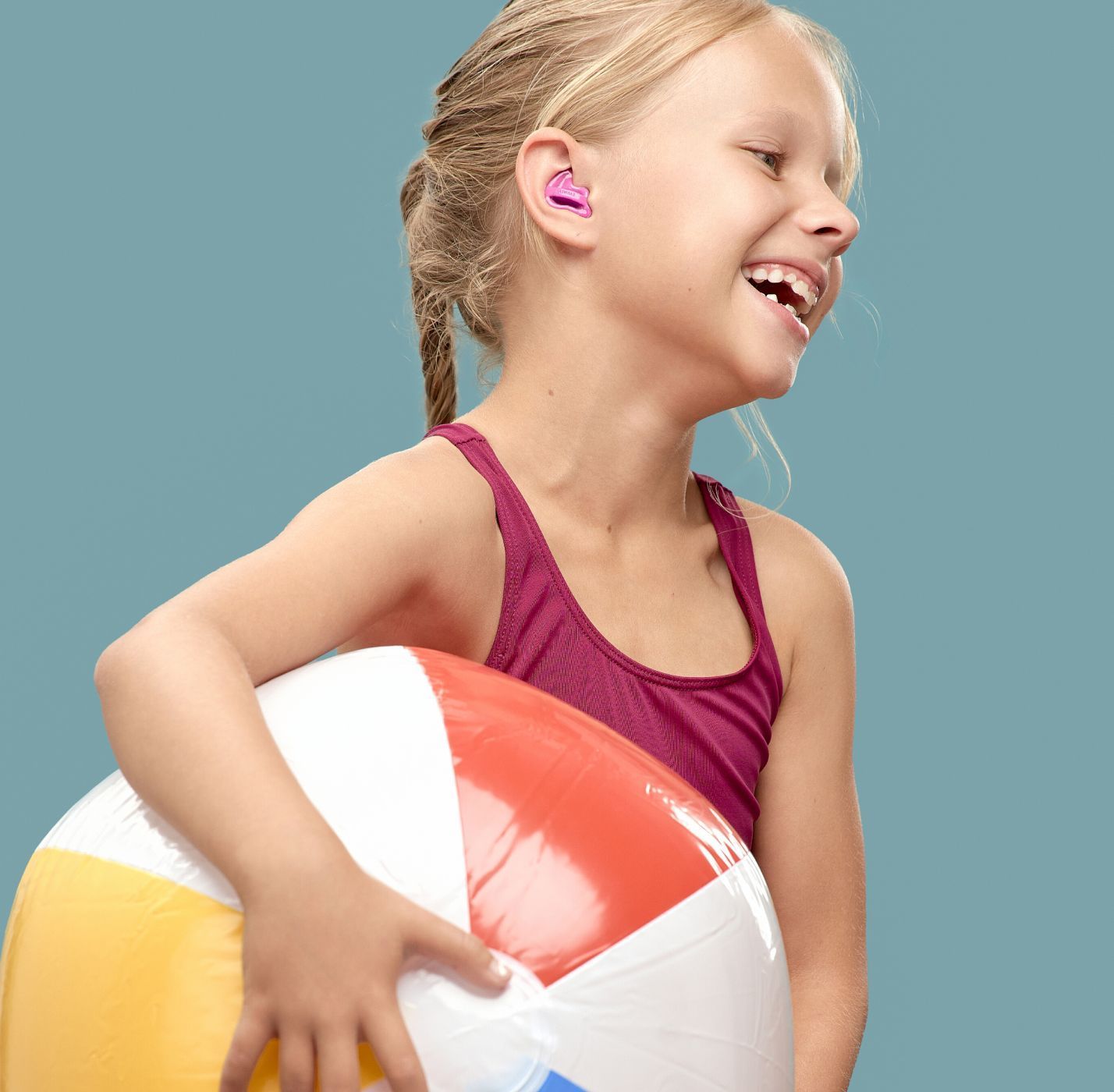 Nasmijana djevojčica u kupaćem kostimu drži loptu za plažu i nosi Earwear Swim zaštitu za plivanje roze boje