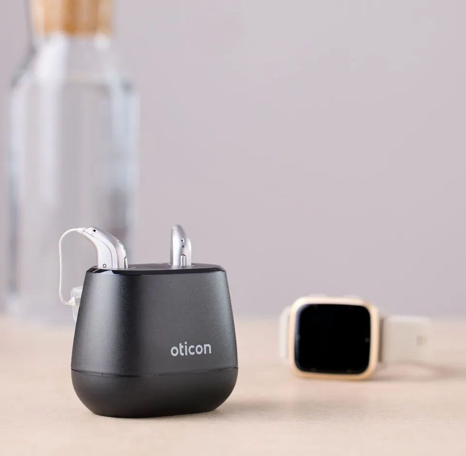 Na mizi stoji polnilna postaja črne barve z napisom Oticon, noter par srebrnih slušnih aparatov, v ozadju je mehko zamegljena steklenica vode in pametna ura s svetlim paščkom in črnim ekranom.