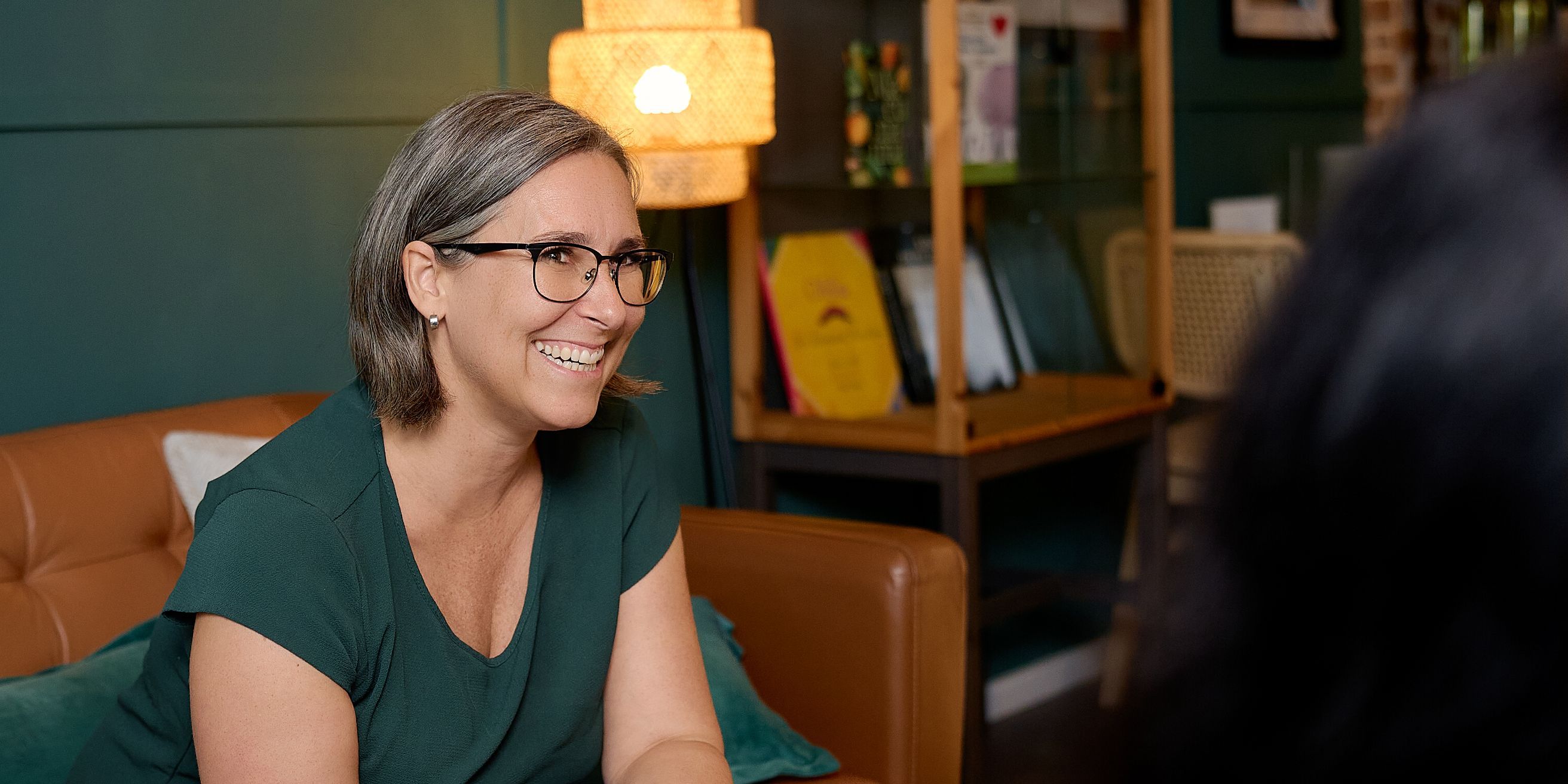 Frau sitzt auf Sofa und lacht während einem Gespräch mit einer anderen Person.