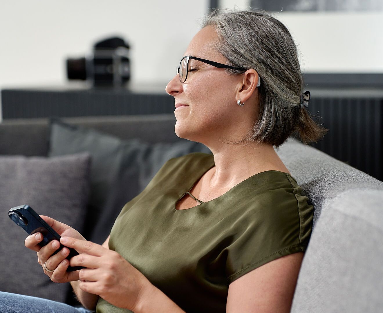 Frau sitzt mit Handy in der Hand auf Sofa und hat ein Hörgerät im Ohr