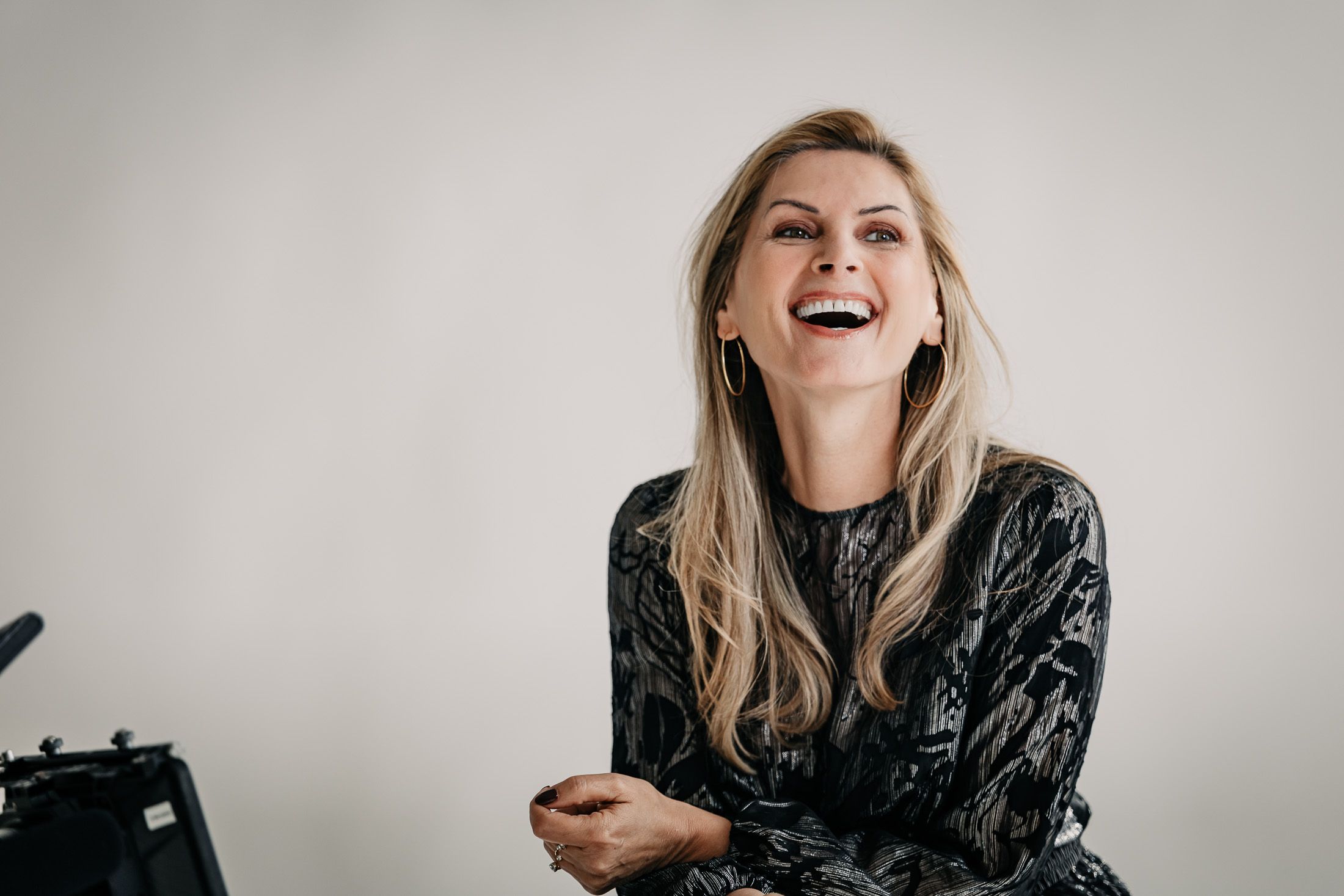 Portrait von lachender Martina Kaiser mit runden Ohrringen in dunklem Kleid vor weißer Wand.