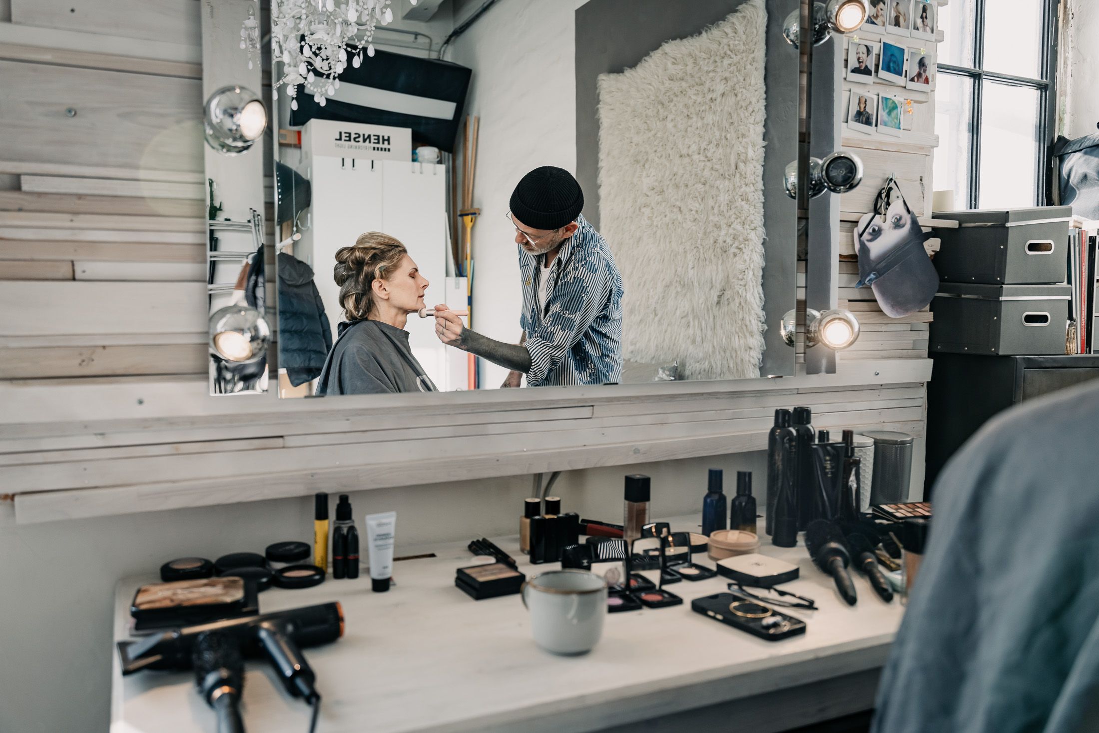 Martina Kaiser vor einem Spiegel in der Maske, Haare in Wicklern und wird von einem Visagisten geschminkt.