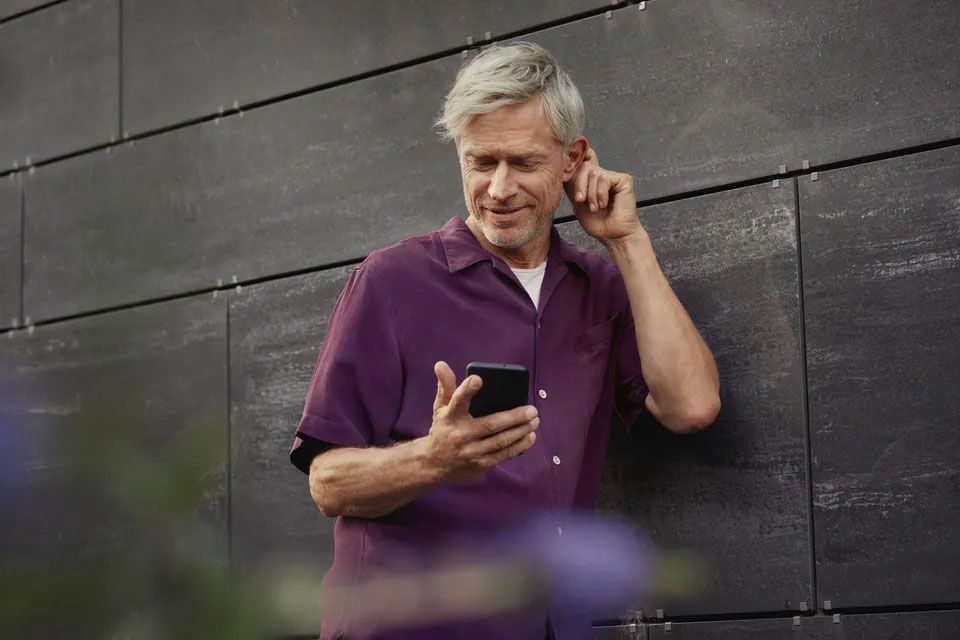 Moški srednjih let s sivimi lasmi, oblečen v vijoličasto srajco kratkega rokava, naslonjen na temno steno, v roki ima pametni mobilni telefon, z drugo roko si nastavlja slušni aparat za ušesom. 