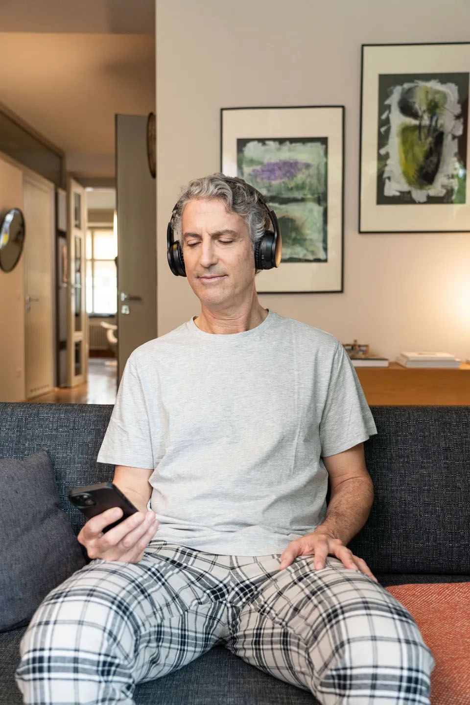 Moški s slušalkami na ušesih, v karirasti pižami sedi na sedežni. V rokah drži telefon. V ozadju ostali del stanovanja - stena s slikami in hodnik, ki pelje v druge prostore. 