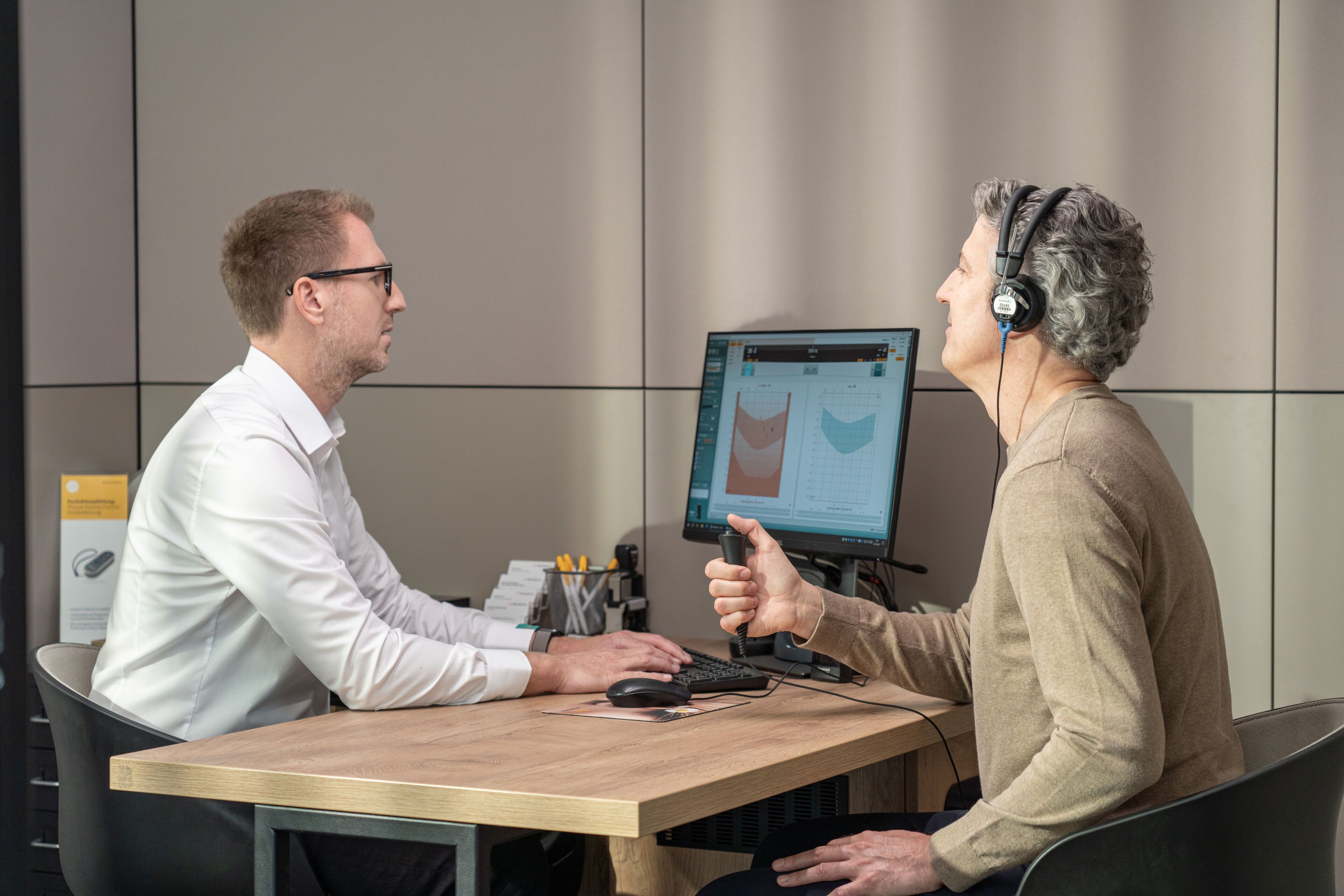 Muškarac sa slušalicama za audiometriju radi slušni test dok ga provjerava akustičar za računalom.