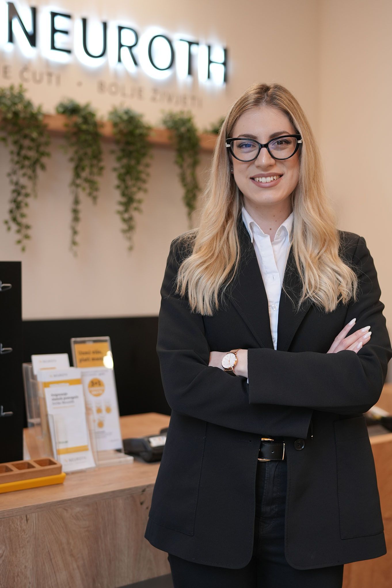 Nasmijana mladda Neuroth akusticarka skrstenih ruku stoji u slusnom centru sa Neuroth logom u pozadini