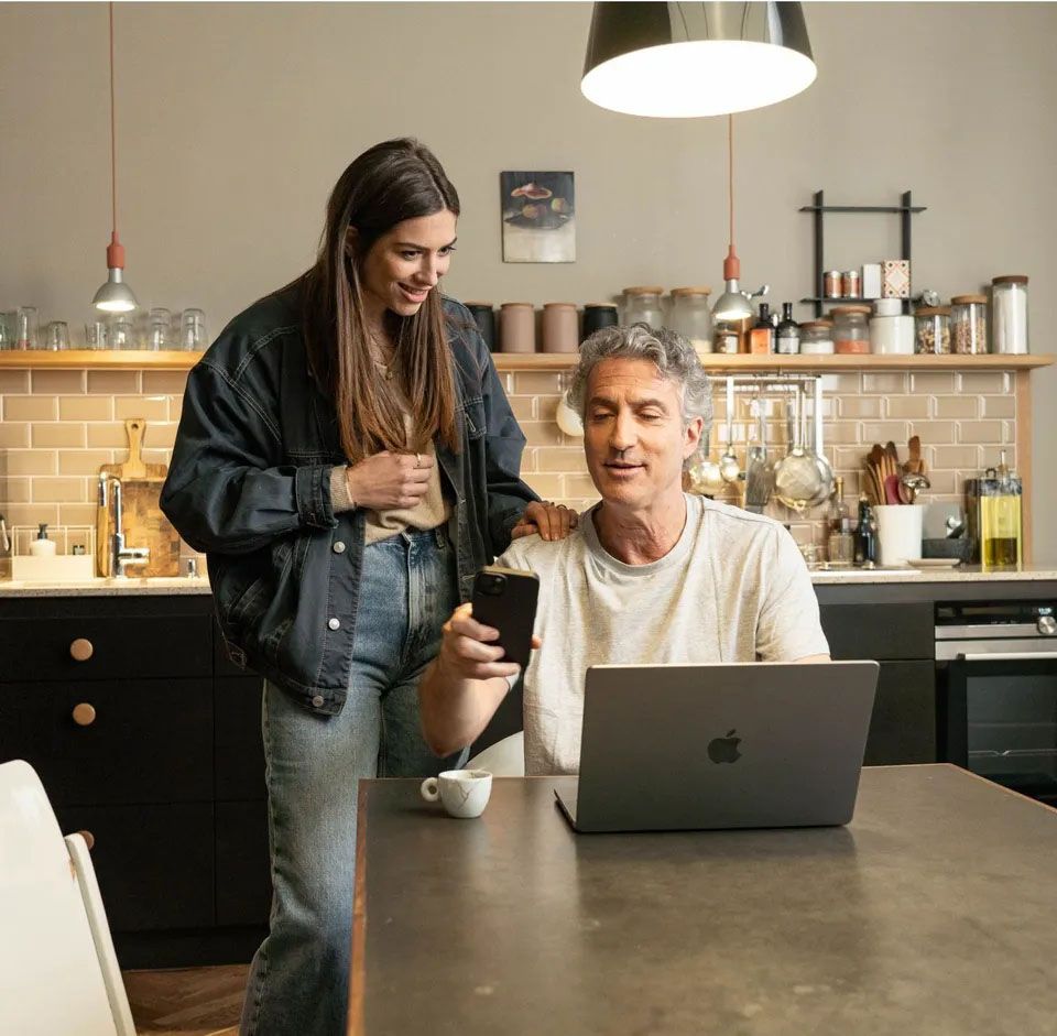 Oče in hči v sodobni kuhinji, oče sedi za mizo s prenosnikom pred seboj in v roki drži pametni telefon, medtem ko hči stoji ob strani in z nasmehom opazuje zaslon, njena roka počiva na njegovem ramenu. 