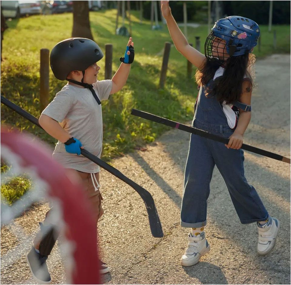 otroci-igrajo-hokejNa sliki sta dva otroka, fant in deklica, ki igrata ulični hokej. Oba nosita zaščitno opremo – fant ima črno čelado in modre rokavice, deklica pa modro hokejsko čelado s kovinsko mrežico. Stojita na tlakovanem igrišču in si z nasmehom izmenjujeta "petko", medtem, ko v rokah držita hokejske palice. V ospredju slike je del hokejskega gola, v ozadju pa se vidi zelena trata in parkirišče.