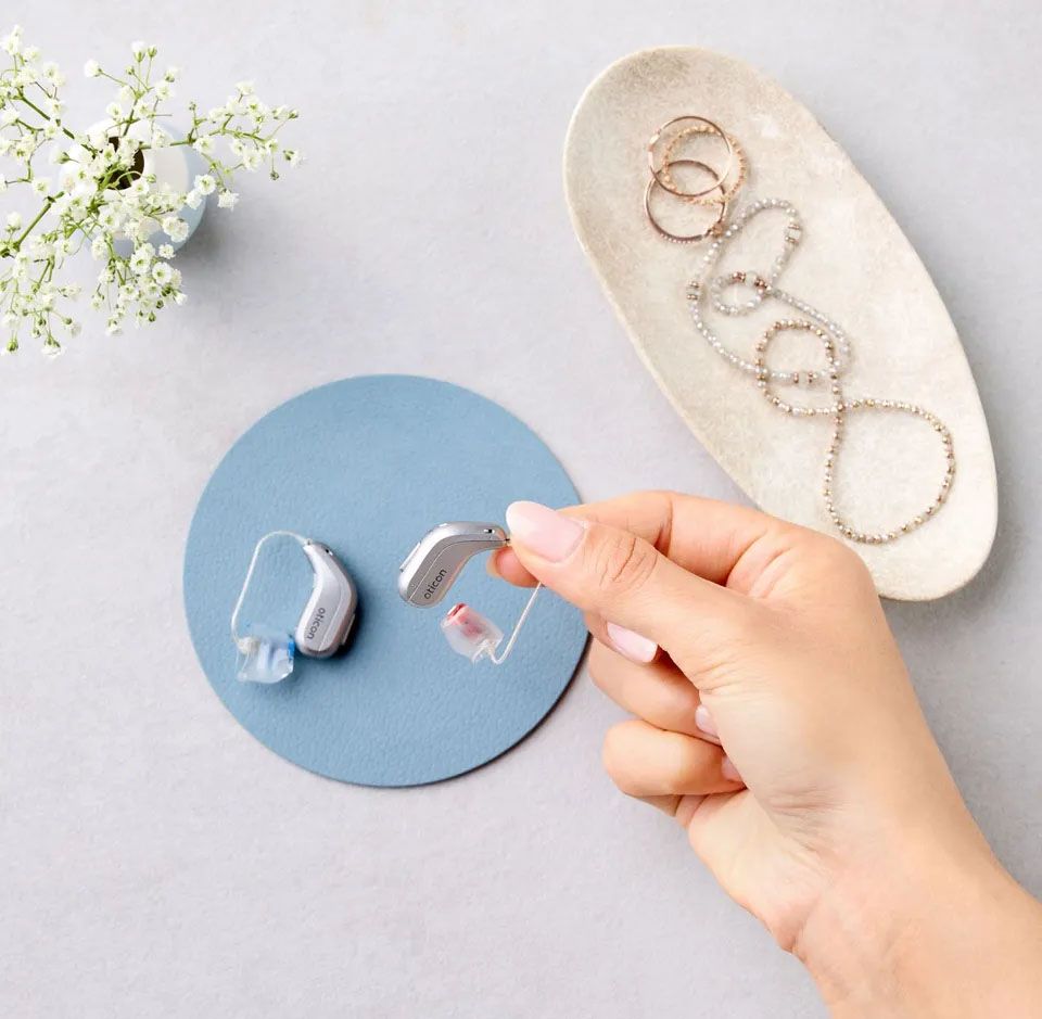 Elegantno urejena scena, ženska roka drži slušni aparat, na podlagi ležita še en enak slušni aparat in krožnik z nakitom, vključno z zapestnicami in prstani bele in zlate barve, ob strani je majhna vaza z rožo z belimi cvetovi.