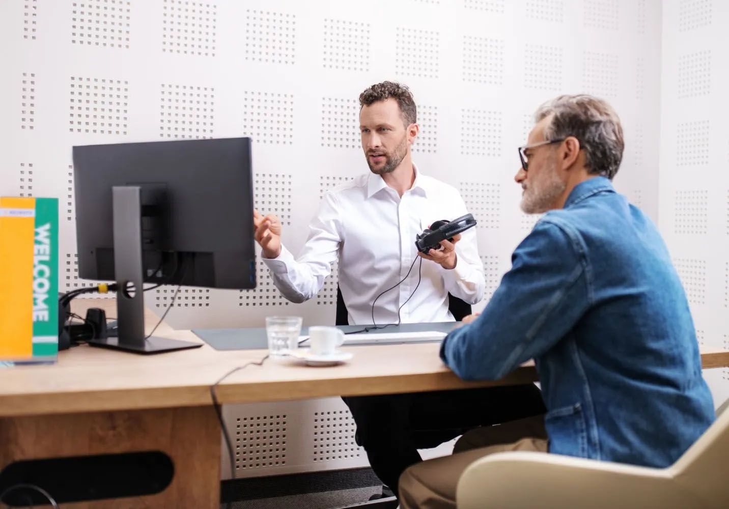Starejši moški na posvetu in testu sluha pri akustičarju. Akustičar v rokah drži slušalke in stranki nekaj razlaga, medtem ko gledata v monitor.