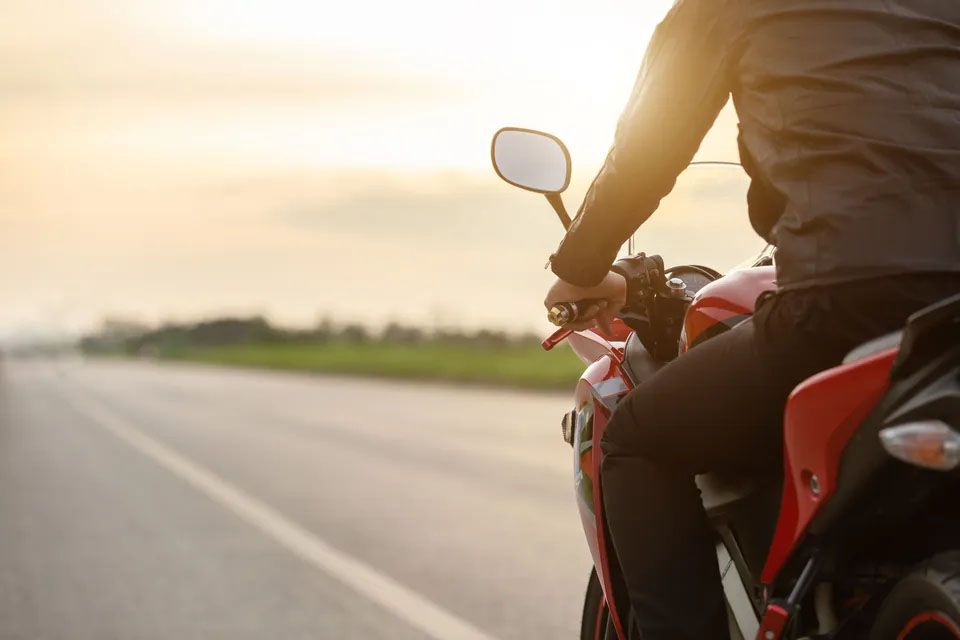 Motorist s črno čelado in rjavo usnjeno jakno na svojem motorju, pelje se bo sivi neoznačeni cesti, okoli posušena trava, zelenje in kamenje, v daljavi se vidi visoka stavba.