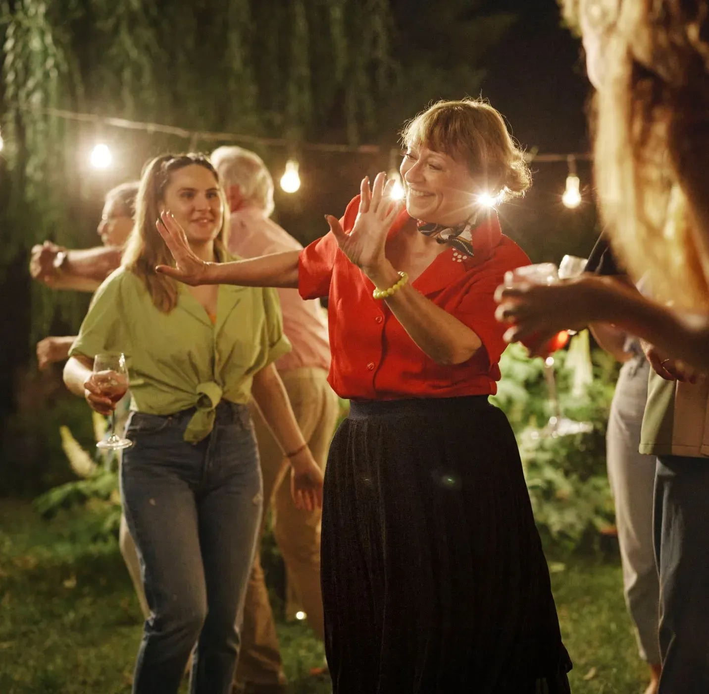 Starija zena u crnoj suknji plese u grupi ljudi u basti na vecernjem partyju