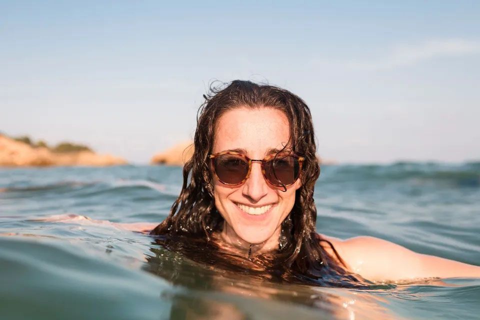 Ženska med plavanjem v morju. Na glavi ima rjava sončna očala, daljši rjavi lasje, v ozadju se vidijo skale. Nasmejana in dobre volje. 
