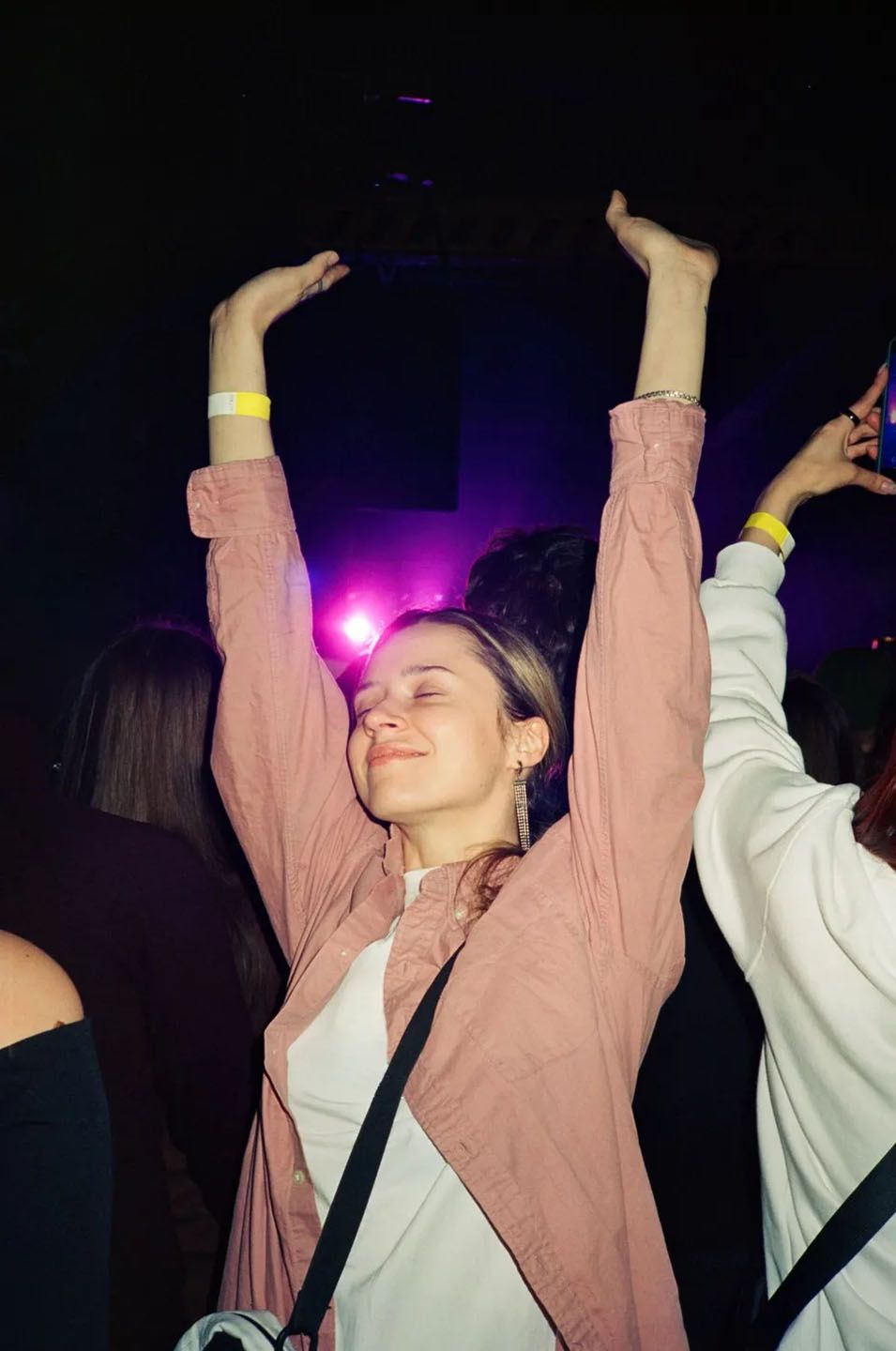 Mlada ženska s svetlejšimi dolgimi lasmi v roza srajci in visečih uhanih, njene roke so dvignjene in oči zaprte, za njeno glavo se blešči roza luč, okoli nje ljude, ki uživajona zabavi.